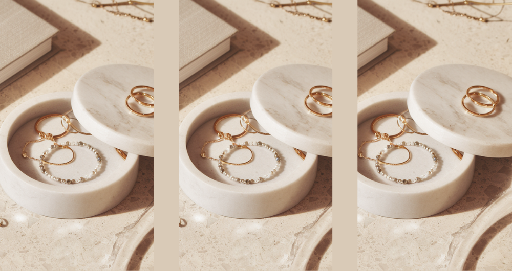 white marble trinket box with a partially removed lid, displaying gold and silver jewelry pieces, including rings, a bracelet, and earrings. The box is placed on a light terrazzo surface with embedded polished stones. A neutral-toned hardcover journal is visible in the background, adding to the elegant and minimal aesthetic