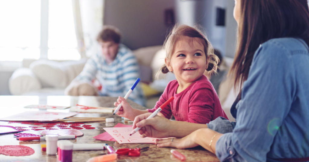 easy valentine’s crafts A cheerful young girl crafting Valentine's Day cards at a table with colorful markers and supplies, smiling at her mom as they create together. A family member relaxes in the background, creating a cozy and heartwarming scene