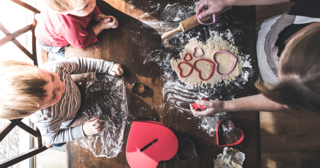 valentine’s day gifts Top-down view of a family baking Valentine's Day cookies together, with heart-shaped cookie cutters and dough on a floured wooden table. Two young children eagerly watch and participate, creating a warm and festive moment