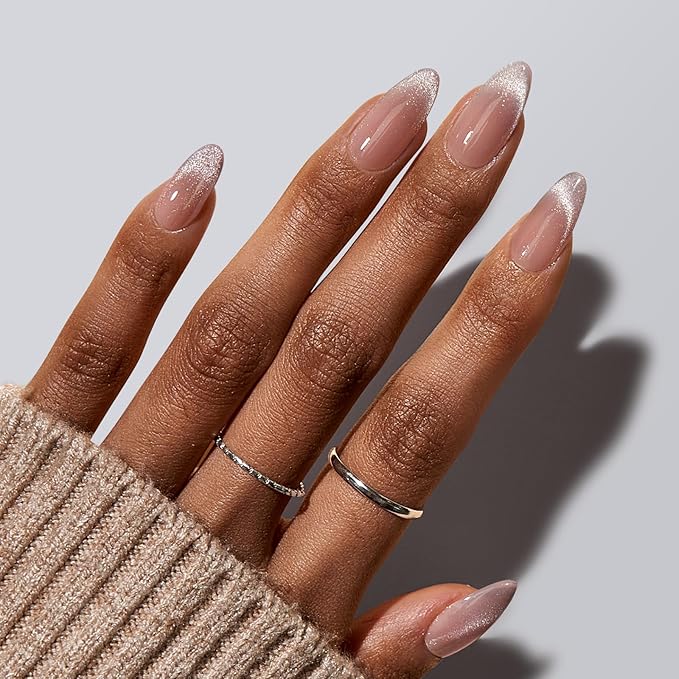 A close-up of a hand with elegant almond-shaped nails featuring a natural pink base and a subtle glittery gradient at the tips. The hand is adorned with minimalistic silver rings, and the person is wearing a cozy beige knit sweater, creating a sophisticated and chic look