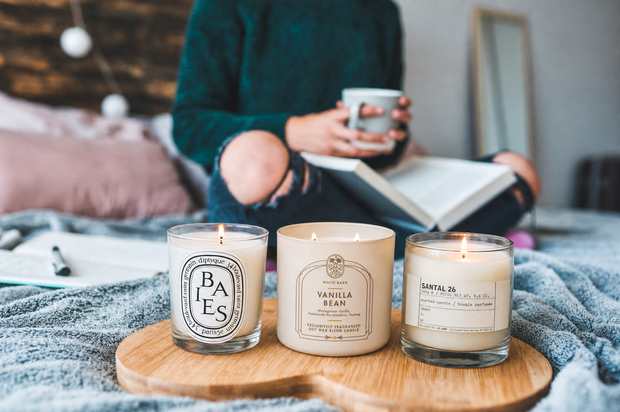 Three luxury candles Diptyque Baies, Bath & Body Works Vanilla Bean, and Le Labo Santal 26—sitting on a wooden heart-shaped tray in a cozy bedroom setting with a woman reading in the background.